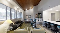 Located at 737 Lockefield Ln in Indianapolis, Indiana, this single-floor space presents a thoughtfully designed interior that blends modern aesthetics with warm, inviting elements. The centerpiece of the space is a dramatic vaulted ceiling finished in rich, natural wood planking, which draws the eye upward and lends the room a sense of grandeur and openness. Recessed lighting is integrated throughout the ceiling, providing ample illumination that complements the natural light streaming in through large, floor-to-ceiling windows along one wall. The main lounge area is furnished with an eclectic yet cohesive mix of seating, including a sleek leather sofa, plush cream-colored sofas, and teal accent chairs arranged around a boldly patterned area rug. Decorative mirrors and carefully curated accessories add character and depth to the walls. A raised platform area at the rear of the space introduces a sense of architectural dimension, featuring a tiled surround and additional seating options. Hardwood-style flooring extends throughout, tying together the various zones of the space with a consistent, polished finish. The overall ambiance is sophisticated yet comfortable, making this an ideal setting for socializing, relaxing, or gathering as a community.