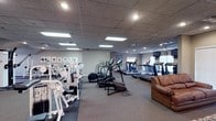This fitness center offers a comprehensive workout environment designed to accommodate a variety of exercise needs. The spacious, single-floor layout is finished with neutral gray walls and a drop-panel ceiling outfitted with recessed and flush-panel lighting that keeps the entire space well-illuminated throughout.

Along one side of the room, a row of treadmills and elliptical machines provides ample cardio options, positioned near a window that brings in natural light. A wall-mounted television is conveniently placed within view of the cardio equipment. On the opposite side, a dedicated strength training zone features a multi-station cable and weight machine setup, ideal for full-body resistance workouts. The flooring transitions between low-pile carpet and rubberized mat sections, appropriately separating the cardio and strength areas.

Motivational artwork and fitness-themed prints line the walls, contributing to an encouraging atmosphere. A leather sofa near the cardio section provides a comfortable spot for rest between sets or for companions waiting during a workout session. The overall ambiance is clean, functional, and welcoming, making this facility well-suited for regular fitness routines across all experience levels.