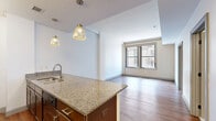 Located at 8 E Long St in the heart of downtown Columbus, Ohio, this one-bedroom, one-bathroom unit offers a well-planned layout with modern finishes across its single floor. The open-concept kitchen and living area serves as the centerpiece of the unit, featuring rich hardwood flooring, a peninsula with granite countertops, pendant lighting, and dark wood cabinetry. The kitchen is equipped with stainless steel and black appliances, including a range, microwave, and refrigerator, with ample counter space on both sides of the galley-style prep area. Large windows throughout the unit frame views of the surrounding downtown Columbus streetscape, allowing natural light to fill the space. The bedroom is a comfortable, well-proportioned room with plush carpeting and oversized windows that continue the urban views. A dedicated closet provides convenient storage. The bathroom features white tile flooring, a granite-topped vanity with dark wood cabinetry, and clean, bright white walls. A utility room rounds out the layout, adding practical functionality. The combination of hardwood and carpet flooring, granite surfaces, and abundant natural light gives the unit a polished, contemporary feel well-suited for urban living.