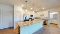 This well-appointed single-floor residence showcases a thoughtful open-concept layout with consistent modern finishes throughout. The kitchen serves as a centerpiece of the space, featuring a generous granite-topped island illuminated by sleek glass pendant lights, crisp white shaker cabinetry, a full suite of stainless steel appliances including a French door refrigerator, microwave, range, and dishwasher, and granite countertops along the perimeter. A distinctive herringbone-patterned hardwood-style floor flows through the kitchen and living areas, adding visual texture and warmth.

The living and bedroom areas continue the clean aesthetic with neutral walls, tray ceilings, and ceiling fans. Multiple bedrooms offer abundant natural light through large windows with outdoor views, and the primary bedroom includes direct access to an outdoor balcony. Storage is well-considered, with louvered closet doors visible in several areas.

The primary bathroom features a dual-sink vanity with granite countertops, a walk-in closet, and a subway tile walk-in shower. A secondary bathroom mirrors the modern design with a subway tile tub-shower combination, granite vanity, and wood-look flooring. Throughout the residence, the bright white palette, clean lines, and consistent material choices create a cohesive, contemporary ambiance.