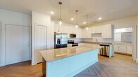 This well-appointed single-floor residence showcases a thoughtful open-concept layout with consistent modern finishes throughout. The kitchen serves as a centerpiece of the space, featuring a generous granite-topped island illuminated by sleek glass pendant lights, crisp white shaker cabinetry, a full suite of stainless steel appliances including a French door refrigerator, microwave, range, and dishwasher, and granite countertops along the perimeter. A distinctive herringbone-patterned hardwood-style floor flows through the kitchen and living areas, adding visual texture and warmth.

The living and bedroom areas continue the clean aesthetic with neutral walls, tray ceilings, and ceiling fans. Multiple bedrooms offer abundant natural light through large windows with outdoor views, and the primary bedroom includes direct access to an outdoor balcony. Storage is well-considered, with louvered closet doors visible in several areas.

The primary bathroom features a dual-sink vanity with granite countertops, a walk-in closet, and a subway tile walk-in shower. A secondary bathroom mirrors the modern design with a subway tile tub-shower combination, granite vanity, and wood-look flooring. Throughout the residence, the bright white palette, clean lines, and consistent material choices create a cohesive, contemporary ambiance.