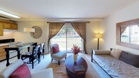 This single-floor unit offers a well-organized layout that balances functionality and comfort across several distinct areas. The living space is anchored by a large sliding glass door dressed with linen-toned draping curtains, allowing generous natural light to pour in while framing views of a landscaped outdoor green space. The open layout accommodates both a seating area and a dining or workspace setup, giving the room a flexible, multifunctional character.

The sleeping area, visible in the dollhouse overview, is integrated within the main living space and features a daybed with a sleek black metal frame and neutral upholstery, complemented by clean white walls and a warm wood-framed mirror. A lean open shelving unit adds practical storage without overwhelming the room.

The kitchen is equipped with warm oak cabinetry extending to the ceiling, a white appliance suite including a refrigerator and range, and a double-basin sink. Woven basket storage atop the cabinets adds a tidy, organized touch.

The bathroom presents a retro-inspired aesthetic with yellow geometric floor tiles, a coordinating toilet and tub, a bold botanical shower curtain, and a wall-mounted shelving unit for storage. A white vanity with cabinetry below completes the space.