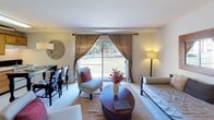 This single-floor unit offers a well-organized layout that balances functionality and comfort across several distinct areas. The living space is anchored by a large sliding glass door dressed with linen-toned draping curtains, allowing generous natural light to pour in while framing views of a landscaped outdoor green space. The open layout accommodates both a seating area and a dining or workspace setup, giving the room a flexible, multifunctional character.

The sleeping area, visible in the dollhouse overview, is integrated within the main living space and features a daybed with a sleek black metal frame and neutral upholstery, complemented by clean white walls and a warm wood-framed mirror. A lean open shelving unit adds practical storage without overwhelming the room.

The kitchen is equipped with warm oak cabinetry extending to the ceiling, a white appliance suite including a refrigerator and range, and a double-basin sink. Woven basket storage atop the cabinets adds a tidy, organized touch.

The bathroom presents a retro-inspired aesthetic with yellow geometric floor tiles, a coordinating toilet and tub, a bold botanical shower curtain, and a wall-mounted shelving unit for storage. A white vanity with cabinetry below completes the space.