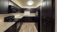 This single-floor space showcases a thoughtfully designed interior centered around a stylish, fully equipped kitchen. The kitchen features rich, dark espresso-toned cabinetry that extends to the upper walls, providing generous storage throughout. Light granite-style countertops offer a striking contrast against the dark cabinetry, creating a bold and contemporary aesthetic. The kitchen is outfitted with a full suite of black appliances, including a refrigerator, electric range with coil burners, built-in microwave, and dishwasher, making it both practical and cohesive in design. A double-basin sink is centrally positioned along the countertop, complemented by a sleek faucet. The U-shaped kitchen layout maximizes workspace and flow, ideal for everyday cooking and meal preparation. Warm wood-look flooring runs throughout, adding a natural, inviting element that complements the darker cabinetry tones. Overhead, a flush-mount ceiling light provides even, ambient illumination throughout the space. The overall design balances functionality with modern style, presenting a clean, well-coordinated interior that is move-in ready.