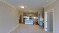 Located on Langdon Road in Morrisville, NC, this single-story space offers a well-designed open-concept layout that integrates the kitchen and living areas into one cohesive environment. The kitchen is a standout feature, showcasing gray shaker-style cabinetry that extends to the upper walls, providing generous storage throughout. A central island with granite countertops serves as both a prep space and a visual anchor for the room, complemented by matching granite surfaces along the perimeter counters. Stainless steel appliances, including a French door refrigerator, microwave, and range, lend a modern, polished look. Pendant lighting hangs above the island, while track lighting illuminates the cooking area, ensuring excellent task lighting. The kitchen flows seamlessly into the carpeted living area, which benefits from the open sightlines and neutral color palette of warm gray tones and white trim throughout. Hardwood-style flooring in the kitchen transitions cleanly to plush carpet in the living space. The overall ambiance is clean, contemporary, and functional, making it a practical and aesthetically cohesive living environment.