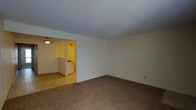 This single-floor residence offers a straightforward and functional layout designed for comfortable everyday living. The main living area is finished with wall-to-wall carpeting, providing a warm and soft underfoot feel, while neutral white walls and a textured ceiling contribute to a clean, understated aesthetic throughout the space. An open passageway connects the living area to the adjacent kitchen, which features tile flooring and is illuminated by overhead pendant lighting that casts a warm glow over the space. The kitchen includes a counter or bar-style divider with white spindle detailing, adding a subtle architectural element to the transition between rooms. A vent is visible near the kitchen entry, indicating a climate control system integrated into the home. The overall ambiance is modest and move-in ready, with a neutral color palette that provides a versatile backdrop for personalization. The single-floor plan ensures ease of navigation throughout the home, making it a practical and accessible living space suited for a range of occupants.