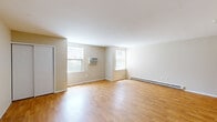 This single-floor unit offers a thoughtfully organized layout comprising a living room, kitchen, bathroom, and closet. The living area is the heart of the space, featuring warm hardwood flooring, neutral beige walls, and clean white ceilings that create a bright, open atmosphere. Multiple windows allow natural light to flow throughout, while a wall-mounted air conditioning unit and baseboard heating ensure year-round comfort. A sliding closet with bifold doors provides convenient storage directly off the living area. The kitchen is efficiently designed with warm honey-toned wood cabinetry, granite-look countertops, a stainless steel sink, and a white electric range with an overhead exhaust hood — offering practical functionality in a compact footprint. The bathroom features a full soaking tub and shower combination, accented by cream-toned wall tiles with a decorative mosaic border in earth tones, lending a clean and polished finish. Throughout the space, consistent hardwood flooring ties the rooms together cohesively. The overall ambiance is clean, neutral, and move-in ready, offering a functional and comfortable living environment.