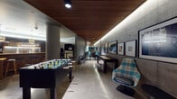 Located at 102 NE 2nd Ave in Portland, Oregon, this single-floor amenity space combines industrial and contemporary design elements to create a multifunctional communal environment. The architecture is defined by striking exposed concrete walls, a warm wood-slatted ceiling, and polished stone tile flooring that runs throughout, lending the space a cohesive and sophisticated character.

A elongated games corridor anchors one side of the space, featuring a foosball table, a shuffleboard table, and plaid upholstered swivel lounge chairs positioned beside small side tables. Framed artwork lines the concrete wall, adding a curated, gallery-like quality to the area. Recessed LED strip lighting runs along the ceiling edge, casting a warm ambient glow that complements the wood slat overhead detail.

Toward the rear, a well-appointed communal kitchen features a long natural wood bar counter with leather-seated bar stools, open warm-toned wood shelving stocked with books and décor, dark cabinetry, stainless steel appliances, and pendant lighting overhead. A wall-mounted television and water dispenser add functional convenience to the area.

An adjacent lounge zone, visible through a frosted glass door, offers casual seating with rounded accent chairs. The overall ambiance is modern and inviting, balancing social activity with relaxed gathering spaces.