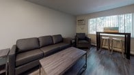 This single-floor unit offers a practical and well-organized layout designed for comfortable everyday living. The open-concept living area is furnished with dark leather seating, a wood-finish coffee table, and a wall-mounted air conditioning unit, providing a cozy and functional space. A window allows natural light to filter in, offering a view of surrounding greenery and contributing to the room's welcoming ambiance.

The kitchen is equipped with grey lower cabinetry paired with white upper cabinets, dark granite-style countertops, and a stainless steel sink. A black range provides a functional cooking station, and the overall design maintains a clean, utilitarian aesthetic consistent throughout the unit.

Hardwood-style flooring runs through the main living and kitchen areas, adding warmth and continuity to the space. The bathroom features a full tub-shower combination with white tile surround, a toilet, and dark countertop cabinetry that mirrors the kitchen's design palette, creating a cohesive look across the unit.

The layout is efficiently organized to maximize use of the available space, making it a straightforward and move-in-ready option. Clean white walls, consistent finishes, and practical fixtures define the overall character of this well-maintained single-floor space.