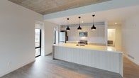 This contemporary single-floor unit showcases a clean, minimalist aesthetic defined by exposed concrete ceilings and light-toned hardwood flooring throughout. The open-concept kitchen is a standout feature, anchored by a generous peninsula island with an illuminated base and sleek light wood cabinetry extending to the ceiling. Three striking black pendant lights hang above the island, adding contrast and visual interest. Stainless steel appliances, including a built-in microwave and refrigerator, complement the modern design, while tall black-framed windows flood the kitchen with natural light and urban views.

The living and bedroom areas continue the unit's bright, airy character, with crisp white walls set against the raw texture of the exposed concrete ceiling. Large black-framed floor-to-ceiling windows offer sweeping city views, enhancing the sense of openness in each room.

The unit features two bathrooms, each finished with neutral large-format floor tiles and clean white surfaces. The primary bathroom includes a double-sink vanity with an integrated white countertop, a wide mirror with integrated storage, and a glass-enclosed walk-in shower with beige subway tile. The second bathroom offers a full soaking tub with a curved chrome shower rod and matching subway tile surround. Together, these spaces deliver a cohesive, contemporary living environment with refined urban style.