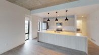 This contemporary single-floor unit showcases a clean, minimalist aesthetic defined by exposed concrete ceilings and light-toned hardwood flooring throughout. The open-concept kitchen is a standout feature, anchored by a generous peninsula island with an illuminated base and sleek light wood cabinetry extending to the ceiling. Three striking black pendant lights hang above the island, adding contrast and visual interest. Stainless steel appliances, including a built-in microwave and refrigerator, complement the modern design, while tall black-framed windows flood the kitchen with natural light and urban views.

The living and bedroom areas continue the unit's bright, airy character, with crisp white walls set against the raw texture of the exposed concrete ceiling. Large black-framed floor-to-ceiling windows offer sweeping city views, enhancing the sense of openness in each room.

The unit features two bathrooms, each finished with neutral large-format floor tiles and clean white surfaces. The primary bathroom includes a double-sink vanity with an integrated white countertop, a wide mirror with integrated storage, and a glass-enclosed walk-in shower with beige subway tile. The second bathroom offers a full soaking tub with a curved chrome shower rod and matching subway tile surround. Together, these spaces deliver a cohesive, contemporary living environment with refined urban style.