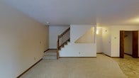 This well-maintained space is organized across three floors, offering a generous and functional layout throughout. The interior showcases a neutral color palette with white walls and warm beige carpeting that runs through the main living areas, creating a clean and understated aesthetic.

A central staircase with warm wood railings and balusters serves as a key architectural feature, connecting the floors with a classic and practical design. The staircase is framed by a solid white partition wall, adding a sense of structure and definition to the open floor plan below.

The lower level living area is notably open and spacious, providing ample room for flexible use. Dark wood baseboards offer a subtle contrast against the light walls, adding a touch of warmth and definition to the rooms. Recessed wall sconce lighting contributes to a soft, ambient glow, complementing the overall neutral tone of the space.

Ceiling heights appear comfortable and consistent throughout, contributing to an airy feel. Ventilation registers are integrated into the walls, indicating a central HVAC system for year-round climate control. The overall condition of the space appears well-kept, with a simple and timeless interior that lends itself to a variety of uses and furnishing styles across all three levels.