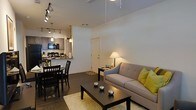 This thoughtfully designed single-floor residence showcases an open-concept floor plan that effortlessly connects the living, dining, and kitchen areas into one cohesive and functional space. The living room is anchored by a tufted gray sofa accented with vibrant yellow-green throw pillows, complemented by a dark wood coffee table resting atop a plush white area rug. Track lighting and a ceiling fan overhead provide both illumination and comfort, while a warm table lamp adds an inviting ambiance to the corner of the room. The dining area features a classic dark wood table and matching chairs, positioned conveniently adjacent to the kitchen. The kitchen is equipped with dark cabinetry, black appliances including a microwave and range, and a breakfast bar that serves as both a functional workspace and casual dining option. Pendant lighting above the bar adds a modern, polished touch. The flooring throughout is a rich dark hardwood, lending warmth and continuity across the open space. The clean white walls and high ceilings contribute to an airy, spacious feel, making this residence both comfortable and visually appealing for everyday living and entertaining.