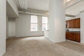 This single-floor unit showcases an industrial-inspired loft aesthetic, characterized by soaring ceilings with exposed ductwork and structural beams that lend a distinctive urban character throughout. The open living area is bathed in natural light from large, tall windows and features plush wall-to-wall carpeting that adds warmth to the expansive space.

The kitchen is thoughtfully designed with rich warm-toned wood cabinetry extending to the upper reaches of the tall walls, complemented by dark granite-style countertops, a checkered tile floor, stainless steel appliances including a side-by-side refrigerator, electric range, and built-in microwave. A peninsula counter provides additional prep space and a casual breakfast bar.

The bedrooms are generously proportioned with high ceilings, soft carpeting, and large windows that allow ample natural light. One bedroom features a ceiling fan, two closets, and views to the exterior. The unit includes two full bathrooms: one with a dual-sink vanity featuring rich wood cabinetry and a separate glass-enclosed shower, and a second bathroom with a matching wood vanity and a full soaking tub. Tile flooring is found in both bathrooms. Overall, the space delivers a bright, airy atmosphere with a cohesive blend of industrial and residential design elements.