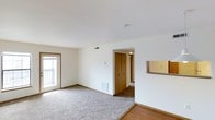 This single-floor residential space offers a clean, functional layout designed for comfortable everyday living. The main living area is finished with plush wall-to-wall carpeting that transitions to hardwood-style flooring near the entryway, creating a warm and welcoming atmosphere throughout. Crisp white walls and standard-height ceilings contribute to a bright, airy feel, while a sliding glass door opens to a private balcony, allowing natural light to fill the room and providing a pleasant outdoor connection. A wall-mounted thermostat and a floor-level heating vent are conveniently positioned in the living area, reflecting practical, well-considered design. A central hallway leads from the living space to the additional rooms of the unit, with a set of bi-fold wooden closet doors providing ample built-in storage directly off the main area. The transition between spaces is seamless, with consistent neutral tones and simple finishes maintained throughout. Overall, the space presents a tidy, move-in-ready environment with a straightforward layout that maximizes usability and comfort.