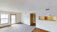 This single-floor residential space offers a clean, functional layout designed for comfortable everyday living. The main living area is finished with plush wall-to-wall carpeting that transitions to hardwood-style flooring near the entryway, creating a warm and welcoming atmosphere throughout. Crisp white walls and standard-height ceilings contribute to a bright, airy feel, while a sliding glass door opens to a private balcony, allowing natural light to fill the room and providing a pleasant outdoor connection. A wall-mounted thermostat and a floor-level heating vent are conveniently positioned in the living area, reflecting practical, well-considered design. A central hallway leads from the living space to the additional rooms of the unit, with a set of bi-fold wooden closet doors providing ample built-in storage directly off the main area. The transition between spaces is seamless, with consistent neutral tones and simple finishes maintained throughout. Overall, the space presents a tidy, move-in-ready environment with a straightforward layout that maximizes usability and comfort.