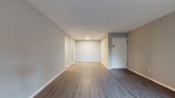 This single-floor unit presents a clean, modern aesthetic characterized by neutral grey and warm beige tones throughout. The open living area is finished with durable laminate flooring in a grey wood-tone, complemented by crisp white baseboards and smooth ceilings with recessed lighting. A sliding window allows natural light to filter into the main living space, which also features a wall-mounted heater unit. A dedicated entry door provides direct access to the unit.

The bedroom is generously proportioned, featuring light-toned laminate flooring, a large window fitted with horizontal blinds that allow for ample natural light, and a closet accessible via a hallway. The hallway connects seamlessly to the other areas of the unit, maintaining an efficient flow throughout.

The kitchen is equipped with white cabinetry, grey laminate countertops, a stainless steel single-basin sink, and stainless steel appliances including a gas range and overhead microwave. Upper and lower cabinet storage is well distributed for practical use.

The bathroom features a full-size soaking tub with a tiled surround, a toilet, and a mirrored medicine cabinet, all finished in clean white tones. Overall, the unit presents a well-maintained, move-in ready interior with a cohesive, contemporary style.