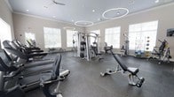 This fitness center offers a thoughtfully designed, single-floor layout that accommodates a comprehensive range of exercise equipment in an open, spacious environment. The main workout area is flooded with natural light from large windows along the walls, creating an inviting and energizing atmosphere. Durable rubber flooring covers the workout space, providing both safety and comfort underfoot.

Overhead, striking circular pendant lights hang from the ceiling alongside ceiling fans, contributing to a modern aesthetic while keeping the space well-ventilated and well-lit. The neutral gray walls and light-toned ceilings keep the environment clean and focused.

The strength training section is centrally anchored by a multi-station cable machine, complemented by a variety of selectorized weight machines, adjustable benches, and free weight equipment. The cardio area features a row of treadmills and additional cardio machines positioned along the perimeter, making efficient use of the available floor space.

A secondary area, visible from the main floor, provides additional equipment and dedicated training zones, including stationary bikes and other specialized machines. The overall layout ensures clear pathways throughout, allowing for safe and comfortable movement between stations. This fitness center is well-suited for both cardio conditioning and full-body strength training.