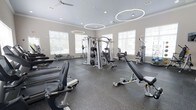 This fitness center offers a thoughtfully designed, single-floor layout that accommodates a comprehensive range of exercise equipment in an open, spacious environment. The main workout area is flooded with natural light from large windows along the walls, creating an inviting and energizing atmosphere. Durable rubber flooring covers the workout space, providing both safety and comfort underfoot.

Overhead, striking circular pendant lights hang from the ceiling alongside ceiling fans, contributing to a modern aesthetic while keeping the space well-ventilated and well-lit. The neutral gray walls and light-toned ceilings keep the environment clean and focused.

The strength training section is centrally anchored by a multi-station cable machine, complemented by a variety of selectorized weight machines, adjustable benches, and free weight equipment. The cardio area features a row of treadmills and additional cardio machines positioned along the perimeter, making efficient use of the available floor space.

A secondary area, visible from the main floor, provides additional equipment and dedicated training zones, including stationary bikes and other specialized machines. The overall layout ensures clear pathways throughout, allowing for safe and comfortable movement between stations. This fitness center is well-suited for both cardio conditioning and full-body strength training.