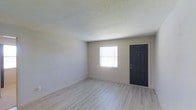 This single-floor space presents a straightforward and functional design with a clean, neutral aesthetic throughout. The main living area features light gray vinyl plank flooring that runs seamlessly across the room, providing a modern and easy-to-maintain surface. The walls are finished in a soft neutral gray tone, contributing to a calm and understated atmosphere that suits a variety of interior styles. The ceiling features a classic textured popcorn finish, adding a familiar residential character to the space. A window fitted with horizontal blinds allows natural light to filter into the room, brightening the interior while maintaining privacy. The bold black front door serves as a distinct architectural accent, creating a strong contrast against the lighter walls and flooring. White baseboards frame the perimeter of the room, adding a crisp finishing detail that ties the overall look together. The open and unobstructed layout of the main area offers flexibility for furniture arrangement and personalization. Overall, the space is well-maintained, move-in ready, and provides a neutral canvas well-suited for a range of uses and design preferences.