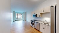 Located at 161 S Huntington Ave in Boston's Jamaica Plain neighborhood, this well-designed one-bedroom, one-bathroom residence offers a thoughtful layout across a single floor. The open-plan kitchen and living area serves as the heart of the home, featuring light hardwood flooring that enhances the bright, airy feel of the space. The kitchen is outfitted with stainless steel appliances — including a range, dishwasher, microwave, and refrigerator — complemented by crisp white shaker cabinetry and granite countertops. Floor-to-ceiling windows along the living area face the street, flooding the space with natural light and offering views of the surrounding neighborhood. The bedroom provides a comfortable retreat with plush carpeting, a large window with outdoor views, and access to a spacious walk-in closet equipped with built-in shelving and additional carpet flooring. The full bathroom features clean tile flooring, a modern vanity, and a well-lit mirror. Neutral tones and clean lines throughout the residence create a contemporary and functional living environment well-suited for urban Boston living.