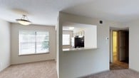 Located at 133 Lake Forest Drive in Daphne, Alabama, this single-story residence offers a well-planned, open layout designed for comfortable everyday living. The living area features neutral-toned carpet flooring and warm beige walls, creating a clean and inviting atmosphere. Large windows with horizontal blinds allow ample natural light to fill the space, with views of surrounding greenery visible from the living area.

A defining architectural feature is the pass-through kitchen opening, which connects the kitchen to the living space via a countertop ledge with a light stone-style surface — ideal for casual dining or entertaining. The kitchen is equipped with stainless steel appliances, including a refrigerator and microwave, complemented by warm wood-toned cabinetry. A window above the kitchen provides additional natural light into the cooking area.

The textured popcorn ceiling and recessed ventilation throughout maintain a consistent, functional aesthetic. Neutral wall colors and carpeted flooring unify the living spaces, giving the unit a cohesive, understated feel. Overall, the space presents a practical and comfortable layout in a quiet residential setting in Daphne, AL.