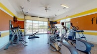 Located at 9200 Bunsen Park Dr in Louisville, Kentucky, this single-level fitness center is designed to support a comprehensive workout experience in an energetic and welcoming environment. The space is distinguished by its bold color scheme, featuring striking orange and yellow horizontal stripe accents along the walls, complemented by a motivational "fit·ness(n.)" graphic that reinforces the room's purpose. Light wood-style flooring runs throughout, adding warmth and contrast to the lively palette.

Natural light is a standout feature, flowing generously through large floor-to-ceiling windows on one side and a full-width glass garage-style door on the opposite wall, which opens to outdoor views of the surrounding community. This combination creates a bright, airy atmosphere that enhances the workout experience. A ceiling fan provides additional airflow and comfort.

The fitness center is thoroughly equipped for both cardio and strength training. Cardio equipment includes multiple treadmills, elliptical machines, a stationary spin bike, and a rowing machine. The strength-training area offers a well-stocked dumbbell rack, kettlebells, a weight bench, an adjustable incline bench, and various exercise balls. Wall-mounted televisions are positioned throughout the space for entertainment during workouts. Full-length mirrors further open up the room visually while supporting proper form during exercise.