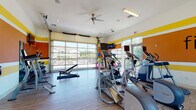 Located at 9200 Bunsen Park Dr in Louisville, Kentucky, this single-level fitness center is designed to support a comprehensive workout experience in an energetic and welcoming environment. The space is distinguished by its bold color scheme, featuring striking orange and yellow horizontal stripe accents along the walls, complemented by a motivational "fit·ness(n.)" graphic that reinforces the room's purpose. Light wood-style flooring runs throughout, adding warmth and contrast to the lively palette.

Natural light is a standout feature, flowing generously through large floor-to-ceiling windows on one side and a full-width glass garage-style door on the opposite wall, which opens to outdoor views of the surrounding community. This combination creates a bright, airy atmosphere that enhances the workout experience. A ceiling fan provides additional airflow and comfort.

The fitness center is thoroughly equipped for both cardio and strength training. Cardio equipment includes multiple treadmills, elliptical machines, a stationary spin bike, and a rowing machine. The strength-training area offers a well-stocked dumbbell rack, kettlebells, a weight bench, an adjustable incline bench, and various exercise balls. Wall-mounted televisions are positioned throughout the space for entertainment during workouts. Full-length mirrors further open up the room visually while supporting proper form during exercise.