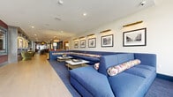 Located in the heart of downtown Columbus, this elegantly designed two-floor space offers a refined and versatile environment spread across a generously sized footprint. The main floor, the larger of the two levels, showcases an expansive lounge area anchored by a sweeping L-shaped blue sectional sofa adorned with floral accent pillows. Brass-accented picture lights illuminate a curated gallery of black-and-white framed photographs, lending a sophisticated, editorial character to the space. Light hardwood flooring runs throughout, complementing the bright, open atmosphere. A stunning wet bar area features dark wood cabinetry, a marble slab backsplash, and open black metal shelving, paired with a curved island and pendant lighting for an upscale, hospitality-inspired feel. A formal dining area with a long white table and coral upholstered chairs adds further functionality. The upper floor presents a more intimate setting, with deep-toned walls, a dramatic chandelier, exposed brick accents near the entry, and cozy wingback chairs flanking a marble-topped coffee table. A semi-private booth nook with textured wallcovering offers a secluded retreat. Large windows on the upper level frame views of the surrounding Columbus skyline, filling the space with natural light.