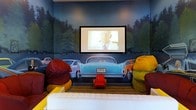 This uniquely designed screening room delivers an immersive, playful take on the classic American drive-in theater experience. The standout feature of the space is a large-scale wall mural depicting a nighttime drive-in scene, complete with painted vintage and modern cars lined up beneath a deep blue, tree-lined sky — creating a dramatic and whimsical backdrop for movie viewing.

A large flat-panel display screen is mounted prominently at the center of the mural wall, positioned to replicate the look of a classic drive-in movie screen. The seating arrangement is equally creative, with an assortment of colorful bean bag chairs in bold shades of red, yellow, and mustard arranged throughout the room, complemented by a low white sofa at the front for additional comfort.

A striped area rug anchors the seating area and adds to the retro, lively aesthetic of the room. The overall color palette — rich blues, deep greens, and vibrant accent colors — contributes to a fun and energetic atmosphere that is both inviting and visually stimulating. The single-floor layout ensures an intimate viewing experience, making this space ideal for casual entertainment and social gatherings centered around film and media enjoyment.
