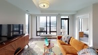 This thoughtfully designed two-bedroom, two-bathroom model residence delivers a comfortable and functional single-floor layout with a contemporary aesthetic throughout. The open-concept living and dining area serves as the heart of the home, featuring hardwood-style flooring, large black-framed windows that invite abundant natural light, and direct access to a private balcony with sweeping urban views. The kitchen flows seamlessly from the main living space and is equipped with stainless steel appliances, including a French door refrigerator and gas range, white shaker cabinetry, and a breakfast bar that provides additional seating and counter space.

The primary bedroom offers a ceiling fan, a generous walk-in closet with built-in shelving and hanging rods, and a large window framing city views. Its en-suite bathroom features a dual-entry layout with a white vanity, tub/shower combination, and a separate glass-enclosed shower stall, complemented by open shelving for storage. The second bedroom is equally well-appointed, with a ceiling fan, carpeted flooring, and convenient access to a dedicated closet. A second full bathroom with a white vanity and light wood-style flooring serves the remainder of the unit. Neutral wall tones and clean finishes create a bright, cohesive ambiance throughout.