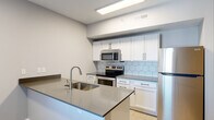 This single-floor unit offers a clean, modern design with thoughtfully selected finishes throughout. The open-concept kitchen serves as the centerpiece of the space, featuring a large central island with a gray quartz countertop and an undermount stainless steel sink. White shaker-style cabinetry with black hardware lines the walls, complemented by a striking hexagonal tile backsplash. A full suite of stainless steel appliances — including a refrigerator, electric range, and over-the-range microwave — completes the kitchen. Light large-format tile flooring runs through the kitchen area, adding to the bright, airy feel of the space. The bedroom is finished with warm wood-look flooring and features a ceiling fan with light fixture, a built-in closet, and a neutral gray color palette that creates a calm, restful atmosphere. The bathroom is elegantly appointed with a dark-framed vanity topped with a white quartz countertop, an oval framed mirror, brushed nickel fixtures, and light tile flooring. Grab bars are also present, reflecting a thoughtful, accessible design. Overall, the unit presents a cohesive, contemporary aesthetic with clean lines and quality finishes throughout.