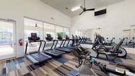 This fitness center is designed to accommodate a wide range of workout needs within a single, open-plan space. The room's most notable architectural feature is its soaring vaulted ceiling, which lends an expansive, airy quality to the interior. Multiple ceiling fans are suspended throughout, complementing the climate control system and ensuring comfortable air circulation during workouts.

Floor-to-ceiling glass windows and doors line one full wall, flooding the space with natural light and offering clear views of the outdoor surroundings. Full-length mirrors are mounted along the opposite wall, visually doubling the sense of space while allowing users to monitor their form. The flooring consists of patterned commercial carpet tiles in a coordinated blue, grey, and tan striped design, providing both durability and visual interest.

The cardio zone is prominently featured and includes multiple treadmills, elliptical machines, upright bikes, spinning bikes, and rowing machines, many equipped with individual digital displays. The strength training area houses a dumbbell rack with a flat bench, a cable/pulley machine, a multi-functional power rack with suspension training attachments, and several selectorized resistance machines. A water fountain and additional functional equipment complete the space. Direct exterior access is available through glass doors on the far end.