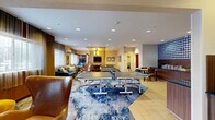 Located at 1383 Washington Ave in Albany, New York, this hotel lobby and social lounge offers a generously proportioned, single-floor space designed to encourage both relaxation and recreation. The open layout flows seamlessly from the front entrance — where a clean, wood-paneled reception desk greets guests alongside a tall fiddle-leaf fig tree — into a vibrant multipurpose lounge area.

The interior is anchored by a bold, abstract blue-and-white area rug that defines the central gathering zone, furnished with a curved leather egg chair, a navy sofa with accent pillows, and sleek side tables. A standout feature is the games area, which includes a full-size billiards table and two ping-pong tables, making the space ideal for social interaction.

Recessed lighting throughout the ceiling provides consistent, warm illumination, while large windows along one wall allow natural light to filter in through sheer curtains. A distinctive accent wall featuring a bold navy polka-dot tile pattern adds a modern, artistic focal point near the reception area.

Additional amenities visible within the space include a wall-mounted television, a small library shelf, and casual dining seating. Light tile flooring in the entry and reception zones transitions smoothly into the carpeted lounge, tying the various functional areas together into one cohesive, welcoming environment.