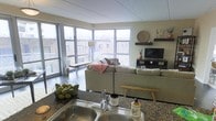 This single-floor space showcases an open-concept layout that integrates the kitchen and living area into one cohesive, functional environment. The most striking architectural feature is the expansive floor-to-ceiling corner windows, which flood the interior with natural light and frame views of the surrounding urban landscape, creating an airy, connected atmosphere throughout.

The kitchen is positioned at the front of the open plan, outfitted with dark granite countertops, a stainless steel sink, and ample counter space suitable for everyday use. The living area beyond is generously proportioned, centered around a large sectional sofa in neutral tones that anchors the room. A dark wood media console supports a flat-screen television, while floating wall shelves display decorative accents, adding character to the space.

The interior palette is kept neutral throughout — warm beige walls, light ceilings, and dark flooring — creating a balanced, contemporary aesthetic. The corner window configuration is a standout design element, maximizing both light penetration and outward views, making the space feel notably larger and more open than its footprint might suggest.