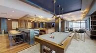This expansive single-floor amenity space blends industrial and contemporary design across several distinct zones. The game room anchors one end of the space, featuring a full-size pool billiards table beneath bold navy-painted ceilings and vintage-style pendant lighting, alongside foosball and additional game options. Exposed brick walls and warm hardwood flooring run throughout, lending an urban, lodge-like character.

A central lounge area hosts a round poker table surrounded by leather chairs, set atop a decorative area rug. Adjacent seating nooks feature reclaimed wood accent walls and soft lounge chairs, creating intimate gathering spots. A café-style dining area with pipe-frame bar tables and navy upholstered seating lines an exposed brick wall, illuminated by industrial sconce lighting.

The communal kitchen is appointed with quartz countertops, warm wood cabinetry with brass hardware, stainless steel appliances, and a patterned tile backsplash. Thick rope details and brick columns add architectural texture nearby.

A sun-drenched reading lounge filled with plush rocking chairs and accent shelving overlooks an outdoor courtyard through a bank of large windows. A sleek mezzanine with glass and steel railings adds vertical dimension, completing this well-appointed communal destination.