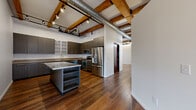 Located at 69 Cascade Dr in Rochester, NY, this 2-bedroom, 3-bathroom residence masterfully blends industrial character with modern finishes across a single thoughtfully designed floor. Rich hardwood flooring runs throughout, complemented by exposed brick walls, original timber ceiling beams, and visible ductwork that lend an authentic loft aesthetic.

The open-concept kitchen and living/dining area serves as the heart of the home. The kitchen is equipped with stainless steel appliances, sleek dark cabinetry, wood-accented countertops, and a central island with built-in shelving. Dramatic track lighting mounted to the exposed beam ceiling adds both function and style. A striking original wood post anchors the space visually.

The generously sized primary bedroom benefits from large windows with outdoor views, exposed brick columns, and wood beam ceilings, creating a warm yet airy retreat. The second bedroom similarly features brick detailing and ample natural light. Three well-appointed bathrooms include wood-toned vanities, glass shower enclosures, and consistent hardwood flooring.

Additional highlights include a balcony, a dedicated closet, a hallway, and a utility area housing HVAC and water heater systems. The bold black entry door and rich mahogany-toned interior doors complete the cohesive industrial-modern design throughout.
