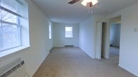 This single-floor space presents a clean, functional layout designed with simplicity and practicality in mind. The main room is generously sized, offering an open and airy feel enhanced by crisp white walls and wall-to-wall carpeting in a neutral beige tone that adds warmth underfoot. Natural light filters in through two windows, lending the space a bright and inviting atmosphere throughout the day. A ceiling fan is centrally mounted overhead, providing both ventilation and supplemental lighting to the room. Baseboard heating units are positioned along the walls, ensuring efficient and consistent climate control during colder months. A doorway visible along one wall leads to an additional area, suggesting further utility or storage beyond the main room. The overall design is straightforward and move-in ready, making it well-suited for a variety of uses. The neutral color palette and uncluttered layout offer a versatile canvas that can easily be personalized to suit individual preferences. This space balances practicality with comfort, making it an accessible and functional option for everyday living.