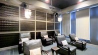 This sophisticated screening room is thoughtfully designed to deliver an immersive cinematic experience within an intimate, upscale setting. The room is anchored by a collection of plush dark leather recliner chairs, each accented with light-toned decorative pillows, arranged to provide every seat with an unobstructed view of the projected screen. Elegant pendant lanterns hang from the dark ceiling, casting a warm ambient glow that complements the room's moody, theater-inspired atmosphere. A ceiling-mounted projector is centrally positioned to deliver a sharp, high-quality projection onto the main viewing wall. The walls are clad in large-format textured acoustic panels with a rich, woven metallic finish, which serve both an aesthetic and functional purpose by enhancing sound quality and minimizing echo. Subtle recessed lighting and wall-mounted speakers are seamlessly integrated into the design, ensuring a clean, polished look throughout. Natural light is softly filtered through shaded windows along one side of the room, allowing for controlled brightness when needed. The overall ambiance is one of refined comfort and purposeful design, making this screening room an ideal venue for private film viewings, presentations, or entertainment gatherings.