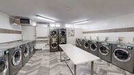 Located at 1340 Danforth Rd in Toronto, this communal laundry facility is a clean, efficiently organized space designed to serve the needs of its users. The room accommodates a generous lineup of commercial-grade front-loading washing machines arranged along two walls, complemented by stacked dryer units that maximize the use of vertical space. A dedicated utility sink provides added convenience for hand-washing or pre-treating garments. At the center of the room sits a large white folding table with a metal frame, offering ample surface area for sorting and folding laundry. The flooring features an eye-catching chevron-patterned tile in soft grey and beige tones, lending a modern aesthetic to the functional space. The walls are finished with a two-tone tile treatment — glossy white subway tiles topped with an accent band of earthy brown tiles — creating a clean and polished look. Overhead fluorescent lighting ensures the room is well-lit throughout. Small windows positioned near the ceiling allow for a degree of natural light and ventilation. An HVAC unit mounted on the wall contributes to climate control within the space. A bulletin board and posted signage provide useful information for users. Security is supported by a visible surveillance camera, and the room is accessible via a standard door.