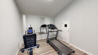 This fitness center in Baton Rouge, Louisiana, offers a clean, dedicated space designed for focused cardiovascular exercise and personal wellness. The room features vaulted ceilings that lend a sense of openness and airiness, while neutral light gray walls create a calm, distraction-free environment conducive to working out. A full-wall mirror installation runs along one side of the room, visually expanding the space and allowing users to monitor their form during exercise. The flooring consists of durable wood-look planks that are both practical and aesthetically polished, complementing the overall modern aesthetic. Natural light enters through a windowed entry door, brightening the space without the need for excessive artificial lighting, though overhead linear lighting ensures visibility at any time of day. A wall-mounted climate control unit keeps the environment comfortable year-round. The fitness center is equipped with a treadmill, a recumbent stationary bike, and an elliptical machine, providing a well-rounded selection of low- to high-impact cardio options. The layout allows for comfortable movement between equipment and leaves additional floor space open for stretching or bodyweight exercises. The space is thoughtfully designed to deliver a functional and welcoming fitness experience.