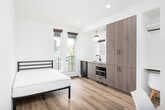 This thoughtfully designed studio presents a clean, modern aesthetic throughout its single-level open-plan layout. Light hardwood flooring and crisp white walls create a bright, airy atmosphere, complemented by recessed lighting and natural light streaming through multiple windows.

The main living area seamlessly integrates the sleeping space with a well-appointed kitchenette. The kitchenette features wood-grain cabinetry in a warm gray-brown tone, a white quartz countertop, a stainless steel undermount sink with a matte black faucet, an induction cooktop, a built-in microwave, and a dishwasher — offering full functionality in a compact footprint. A pendant globe light adds a stylish accent above the kitchen area. French doors open to a balcony, flooding the space with natural light and providing pleasant outdoor views.

A dedicated workspace nook includes a built-in desk and seating, offering a practical area for studying or working. An open closet with a hanging rod provides convenient wardrobe storage nearby.

The private bathroom is finished in the same light, contemporary palette, featuring a vanity with wood-tone cabinetry, a toilet, and a separate walk-in shower stall with bright white surround panels and brushed nickel fixtures. A window in the bathroom adds natural ventilation and light. The overall design is efficient, cohesive, and move-in ready.