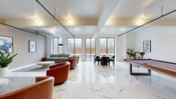 This contemporary amenity lounge is a thoughtfully designed, open-concept space that seamlessly blends comfort and entertainment. The floor is finished in large-format polished marble tiles with striking veined detailing, lending an air of sophistication throughout. Structural beams and industrial-style track lighting run across the ceiling, adding an architectural contrast to the otherwise clean, white interior.

The space is organized into several distinct functional zones. A lounge area features plush grey sofas, curved cognac leather swivel chairs, and low round coffee tables arranged on a textured area rug, creating an inviting setting for relaxed conversation. A wall-mounted flat-screen TV and mounted abstract artwork contribute to the curated aesthetic. An additional seating nook near the windows offers a quieter retreat with a grey sofa and warm wood side tables.

A billiards table with a rich walnut finish serves as a central entertainment focal point, positioned beneath the coffered ceiling detail. A round dining table and sleek black chairs provide a dedicated space for gathering. Throughout the room, expansive black-framed floor-to-ceiling windows flood the interior with natural light and frame views of the outdoor surroundings, enhancing the open, airy ambiance of the entire space.