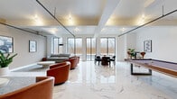 This contemporary amenity lounge is a thoughtfully designed, open-concept space that seamlessly blends comfort and entertainment. The floor is finished in large-format polished marble tiles with striking veined detailing, lending an air of sophistication throughout. Structural beams and industrial-style track lighting run across the ceiling, adding an architectural contrast to the otherwise clean, white interior.

The space is organized into several distinct functional zones. A lounge area features plush grey sofas, curved cognac leather swivel chairs, and low round coffee tables arranged on a textured area rug, creating an inviting setting for relaxed conversation. A wall-mounted flat-screen TV and mounted abstract artwork contribute to the curated aesthetic. An additional seating nook near the windows offers a quieter retreat with a grey sofa and warm wood side tables.

A billiards table with a rich walnut finish serves as a central entertainment focal point, positioned beneath the coffered ceiling detail. A round dining table and sleek black chairs provide a dedicated space for gathering. Throughout the room, expansive black-framed floor-to-ceiling windows flood the interior with natural light and frame views of the outdoor surroundings, enhancing the open, airy ambiance of the entire space.