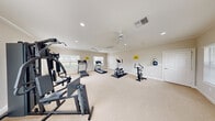 The Sundance Apartments Gymnasium is a dedicated fitness facility offering a comfortable and functional workout environment for residents. The single-room exercise space is finished with neutral-toned carpet flooring and light beige walls, creating a clean and inviting atmosphere throughout.

The room is brightened by multiple windows along the walls, allowing natural light to flow in and providing views of the surrounding community. Dual ceiling fans with recessed lighting ensure proper air circulation and illumination, keeping the space comfortable during workouts.

Crown molding along the ceiling adds a refined architectural touch, while the open floor plan provides ample room to move freely between stations. Equipment includes treadmills, an elliptical cross-trainer, an upright exercise bike, and a multi-station cable strength-training machine, offering a well-rounded selection for both cardio and resistance training.

Wall-mounted flat-screen televisions are positioned for easy viewing, and a water dispenser is conveniently available near the wall. Safety signage is posted throughout the room, and the space also features a storage closet with double doors. A dedicated entry door with nearby wall-mounted controls completes the practical layout of this community fitness amenity.