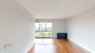 This single-floor unit offers a clean, minimalist layout designed for comfortable everyday living. The main living area is a standout feature, centered around a large picture window that fills the room with abundant natural light and frames sweeping views of lush treetops and the surrounding cityscape beyond. Warm-toned hardwood flooring runs throughout, adding a sense of continuity and warmth to the neutral, light-grey walls and white trim. The textured ceiling adds a subtle classic character to the space.

The kitchen is efficiently laid out in a galley style, featuring rich dark-wood cabinetry, a double-basin sink, a gas range, and a full-size refrigerator — all finished in black for a cohesive look. A window in the kitchen provides additional natural light, enhancing the functionality of the space.

The bedroom area is equipped with a built-in closet organizer system, complete with multiple hanging rails and integrated shelving, offering practical and ample storage. The entry area flows naturally into the main living space, maintaining an open and unobstructed feel throughout. Overall, this unit presents a well-maintained, light-filled interior with practical finishes and pleasant outdoor views.
