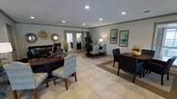 This single-floor professional space is designed to be both functional and inviting, offering a polished environment suited for client-facing operations. The main area serves as a combined reception and workspace, featuring light-toned large-format tile flooring that enhances the sense of openness throughout. Multiple workstations are thoughtfully arranged to accommodate staff and visitors, each furnished with rich dark wood desks paired with upholstered accent chairs in soft, neutral tones. Recessed lighting is installed throughout the ceiling, providing consistent, warm illumination across the space. Decorative touches include a set of ornate sunburst mirrors, framed abstract wall art in muted greens and blues, and potted tropical plants that add a refreshing natural element. A set of glass-paneled French doors at the rear of the space allows natural light to filter in and provides a visual connection to an adjoining area or exterior. An area rug anchors one of the seating clusters, adding texture and definition to the layout. The overall ambiance is professional yet welcoming, with a carefully curated aesthetic that balances practicality with comfort, making it an ideal setting for consultations or administrative operations.
