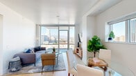 This contemporary urban residence is designed with clean lines, a neutral palette, and an abundance of natural light. The open-concept living and dining area is anchored by expansive floor-to-ceiling windows and sliding glass doors that frame striking panoramic views of the city skyline and surrounding mountains, creating a bright and airy atmosphere throughout. Light hardwood flooring flows seamlessly across the main living spaces, complementing the crisp white walls and modern aesthetic.

The kitchen is thoughtfully appointed with high-gloss white upper cabinetry, warm wood lower cabinets, a subway tile backsplash, and stainless steel appliances including a gas range, microwave, and dishwasher. A dedicated dining area sits adjacent to the kitchen, offering a natural transition between cooking and entertaining spaces.

The bedroom is a tranquil retreat bathed in natural light, featuring large windows and easy access to additional storage. Two well-appointed bathrooms serve the residence — each finished with large-format tile, wood-toned vanities, white countertops, and a bathtub with glass enclosure. A dedicated laundry closet with stacked units adds everyday convenience. Recessed lighting throughout ensures a polished, consistent finish across every room.