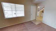 This two-story residence offers a straightforward and functional layout designed for everyday living. The main level greets visitors with a tiled entryway that transitions seamlessly into a carpeted living area, creating a clear distinction between spaces. A large window fitted with horizontal blinds allows natural light to fill the room, brightening the neutral-toned walls and contributing to an open, airy atmosphere. The front door opens directly into this welcoming zone, making for a practical and accessible entry point. Adjacent to the entry, a staircase with wooden steps and a light-toned railing connects the main level to the upper floor, adding vertical dimension to the home. The open sightline from the living area through to the staircase and beyond gives the space a sense of flow and continuity between levels. The neutral color palette throughout — from the beige walls to the light carpet — provides a clean, versatile backdrop well-suited to a variety of furnishing styles. Overall, this two-floor residence balances practicality with a comfortable, livable ambiance, making it an approachable and adaptable home.
