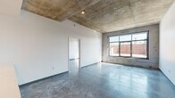 Located at 121 Delaware St in Kansas City's vibrant River Market neighborhood, this single-floor unit showcases an industrial-modern aesthetic defined by exposed concrete ceilings and polished concrete floors throughout. The open layout connects the living area to a well-appointed kitchen featuring white upper cabinetry, warm wood-toned lower cabinets, light quartz countertops, stainless steel appliances including a refrigerator, range, and microwave, and a central island with pendant lighting overhead. The exposed concrete ceiling with structural beams adds architectural character to the kitchen and living areas alike. Large, black-framed windows in the living and bedroom spaces flood the interior with natural light while framing views of Kansas City's surrounding urban landscape, including glimpses of the downtown skyline. The bedroom benefits from its own generously sized window with similar city views. The bathroom is finished with a warm wood vanity, white quartz countertop, full-width mirror, and a tiled tub-shower combination with a handheld showerhead. Polished concrete floors continue seamlessly into the bathroom, reinforcing the cohesive industrial design language found throughout the entire unit.